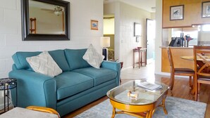 A living room with a teal sofa, two patterned pillows, a glass coffee table, a wooden side table, and a dining area with wooden chairs and a table. - KAILANI at Mauna Loa Shores, Fantastic Condo, Swim with the Sea Turtles (Hilo)