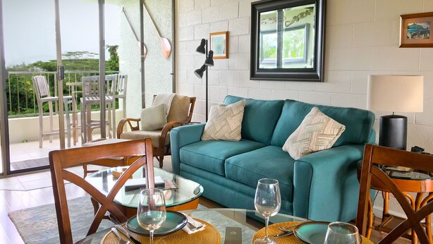 A teal sofa with patterned cushions, a glass table with wine glasses, and a view of greenery through a glass door. - KAILANI at Mauna Loa Shores, Fantastic Condo, Swim with the Sea Turtles (Hilo)