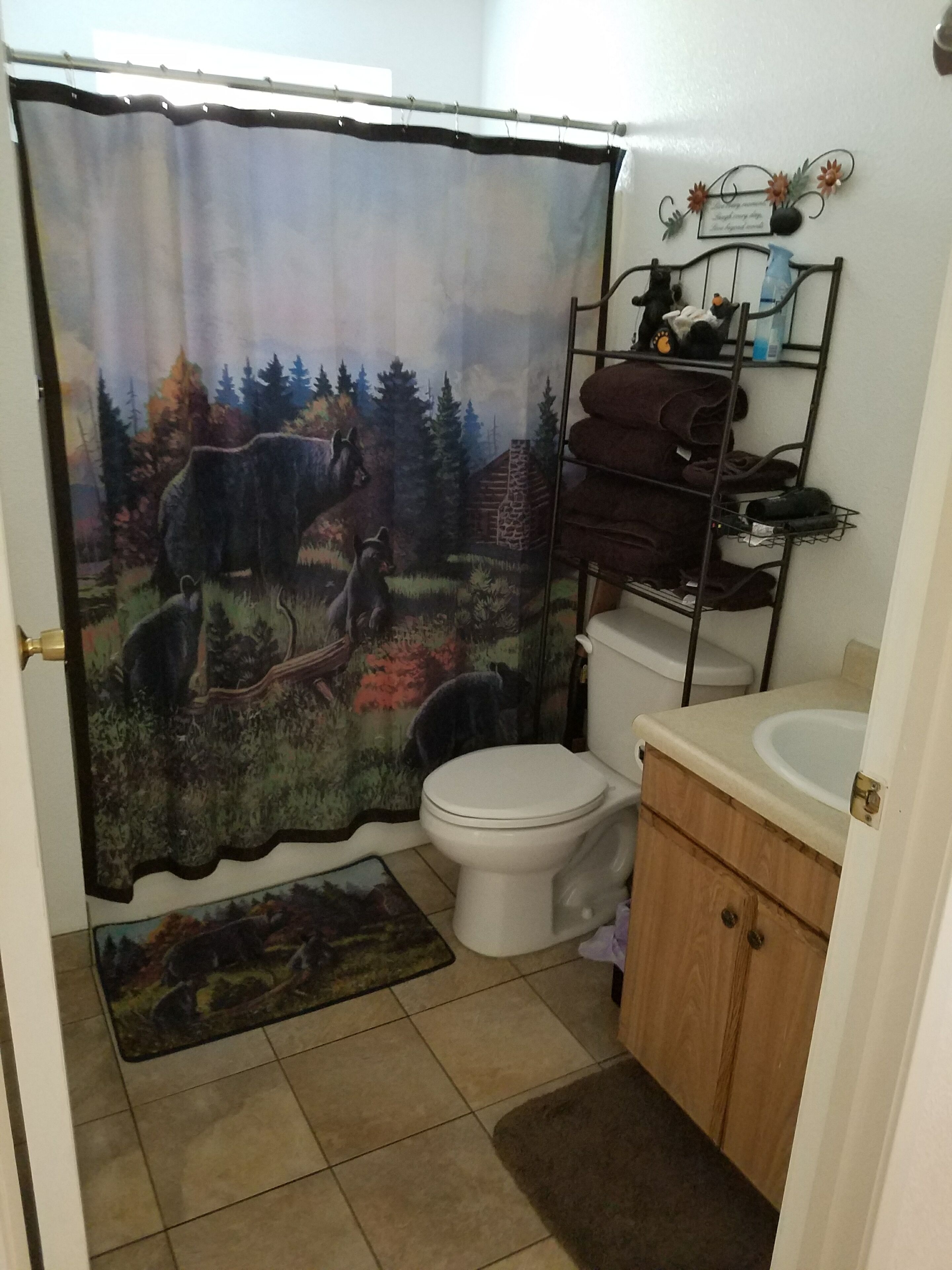 Un baño con cortina de ducha que muestra un paisaje forestal, un inodoro, un lavabo y un perchero para toallas.