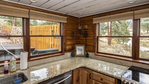 A kitchen with granite countertops, wooden cabinets, a stainless steel dishwasher, a stove, and large windows with a view of a wooden fence and trees.