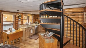 A log cabin interior with a spiral staircase, wooden furniture, a kitchen area, and a dining table.