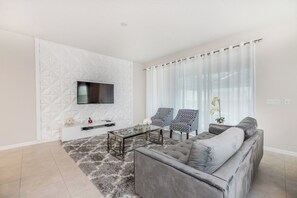 Un salon moderne avec un canapé gris, deux fauteuils à motifs, une table basse en verre et un téléviseur à écran plat fixé à un mur texturé.