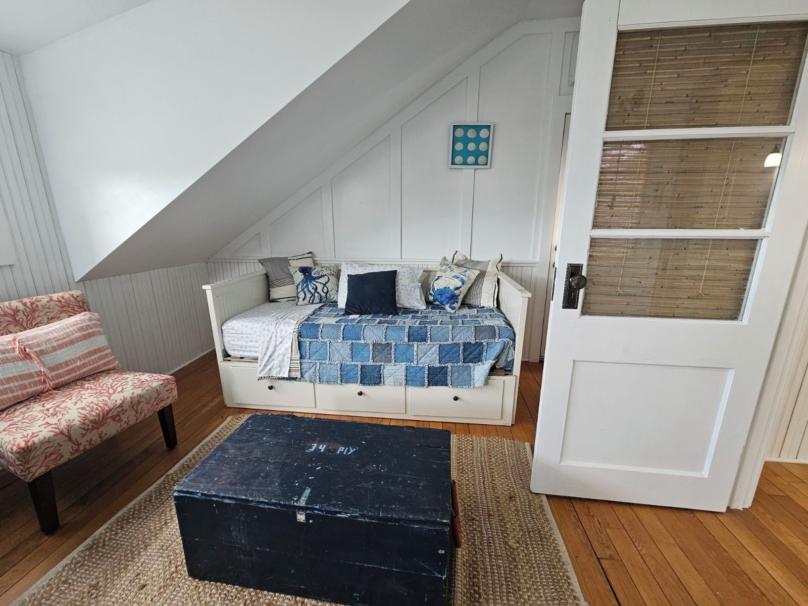 A loft bedroom with a sloped ceiling, a bed with a blue quilt, a chair with a floral pattern, and a black chest of drawers.