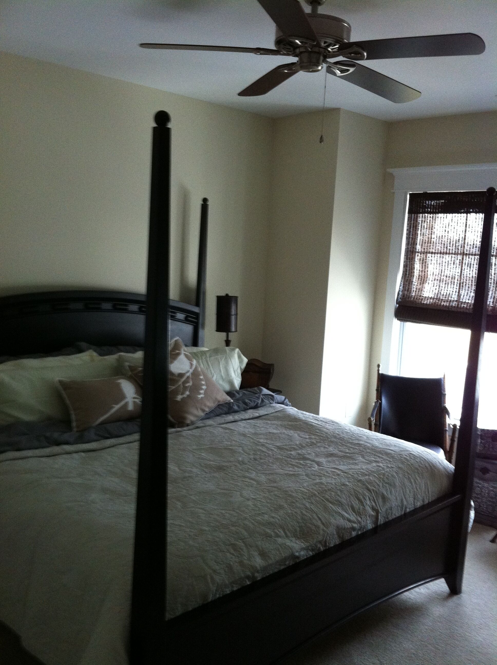A four-poster bed with a ceiling fan, a window with blinds, and a nightstand with a lamp.