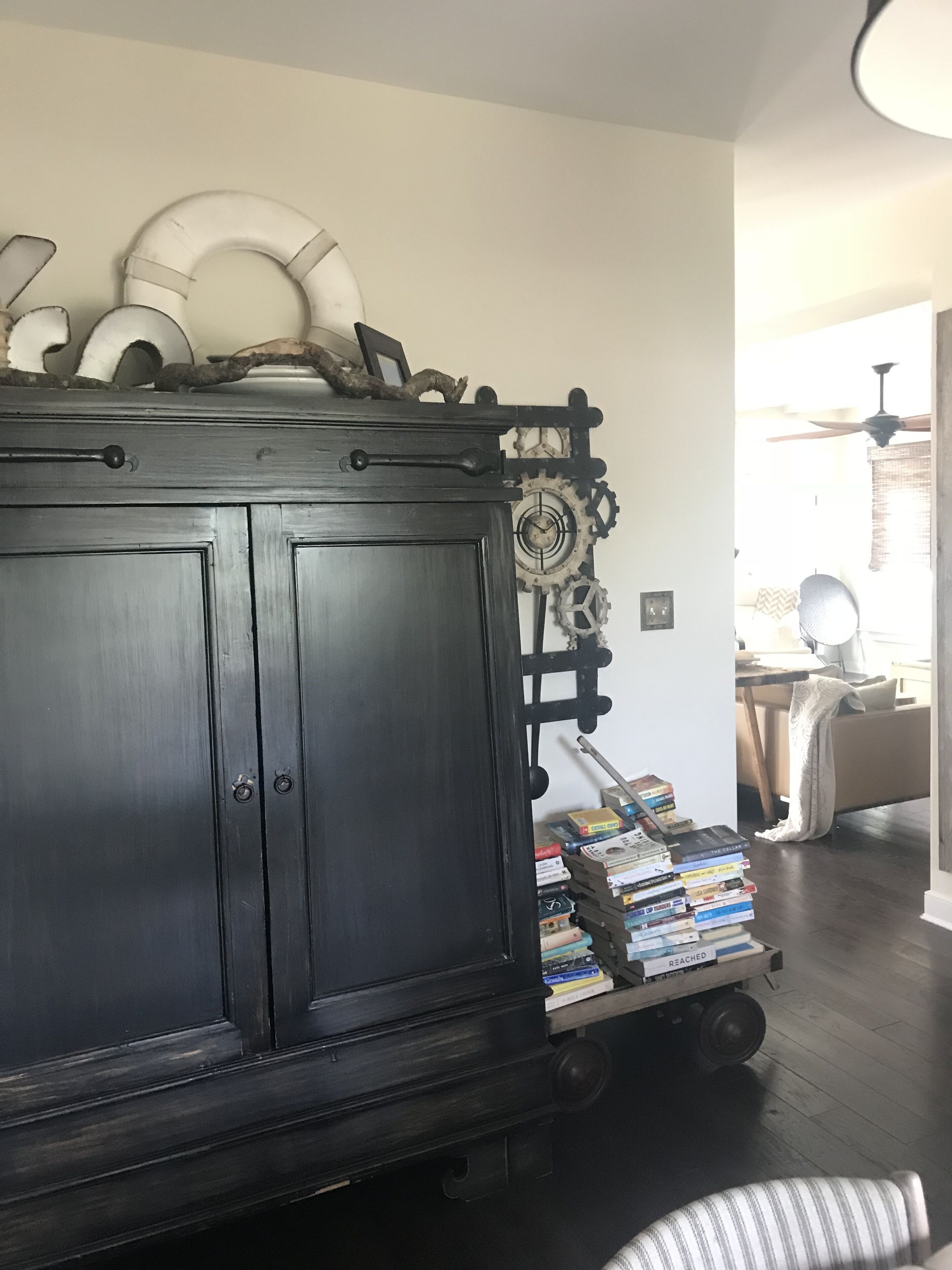 A dark wooden cabinet with a clock and decorative gears, a small cart with books, and a ceiling fan in a room with a light-colored wall and wooden floor.