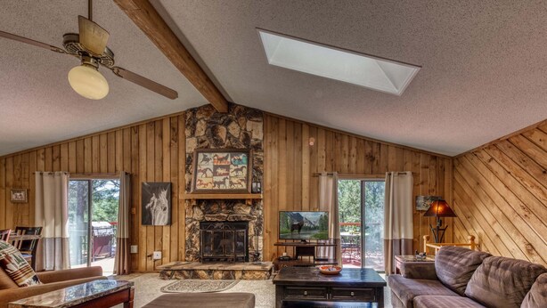 A log cabin interior with a stone fireplace, wooden walls, a ceiling fan, and a television.