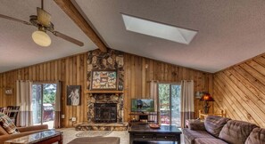 A log cabin interior with a stone fireplace, wooden walls, a ceiling fan, and a television.