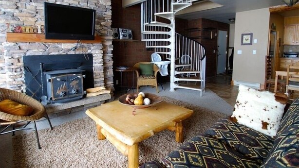 Una sala de estar acogedora con pared de piedra, chimenea, mesa de centro de madera, sofá y una escalera espiral que lleva a otra habitación.
