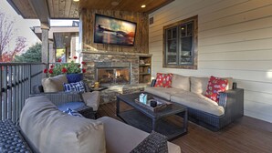 A covered outdoor seating area with wicker furniture, a stone fireplace, and a flat-screen TV mounted on the wall. - Incredible luxury townhome in downtown Whitefish! 1 block from all downtown shops and restaurants (Whitefish)