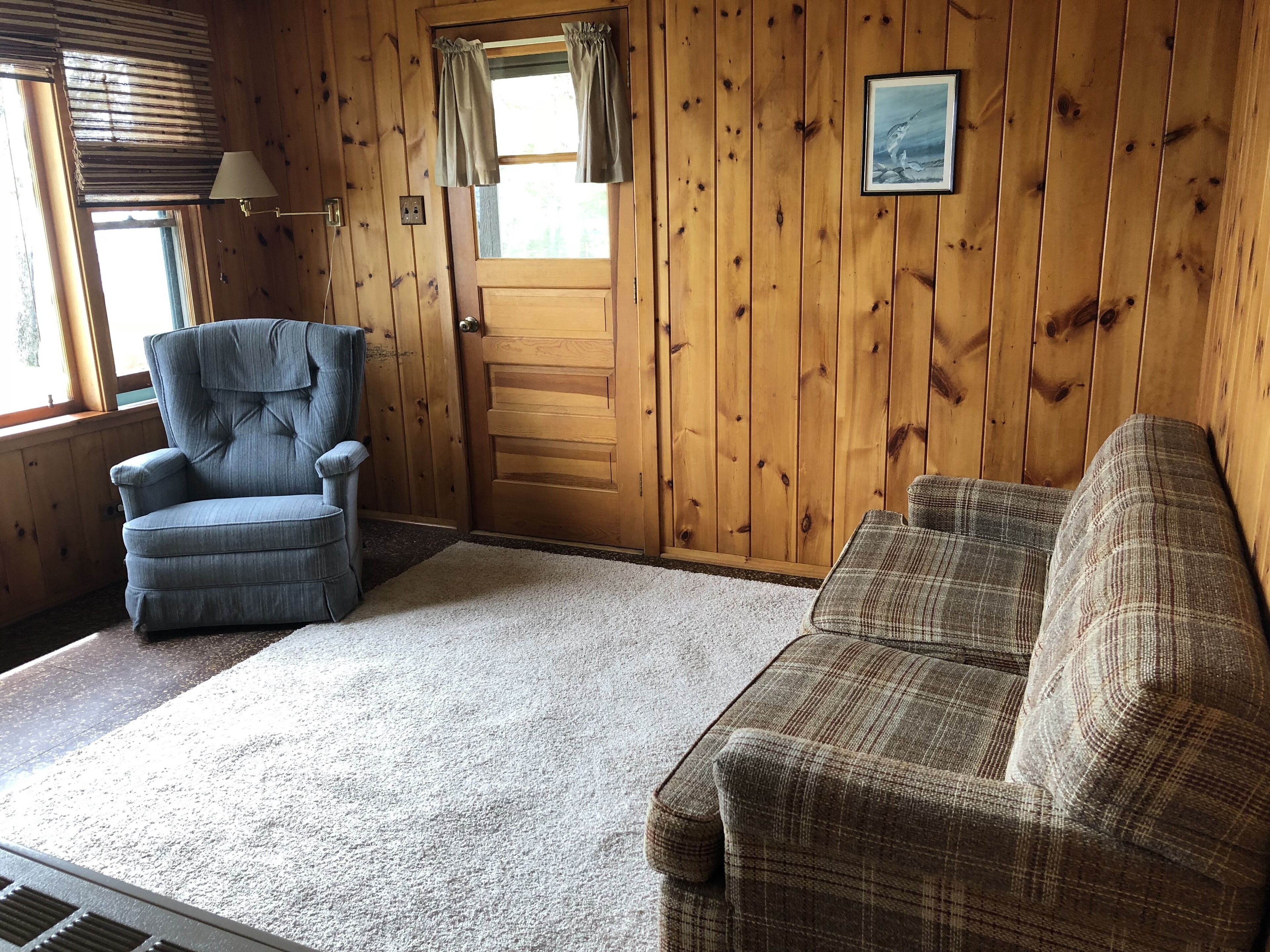 A room with wooden walls, a blue armchair, a plaid sofa, a door, and a window with curtains.