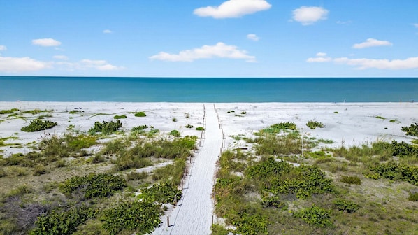 Une plage de sable avec un étroit sentier menant à l’océan, bordée d’une végétation clairsemée.