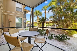 A balcony with wicker chairs and a table, overlooking a palm tree garden and a building with a wooden facade.