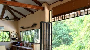 Zona pranzo con tavolo in legno apparecchiato per quattro persone, circondato da sedie in vimini con cuscini rossi. In secondo piano si vede una cucina con piano cottura e utensili.