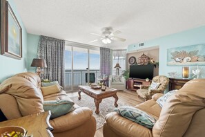 A living room with leather sofas, a coffee table, a TV, and a balcony with curtains. - Upscale Refined Unit, Oceanfront Water Amenities | Crescent Shores - N 1304 (North Myrtle Beach)