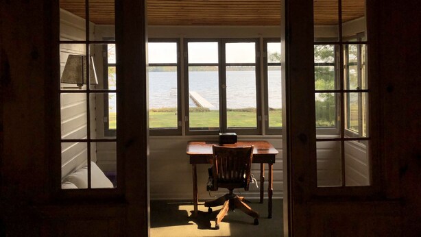 A room with a desk and chair facing a large window overlooking a body of water. - Where eagles soar, the sun sets low and the deer are friendly.  (Saint Germain)