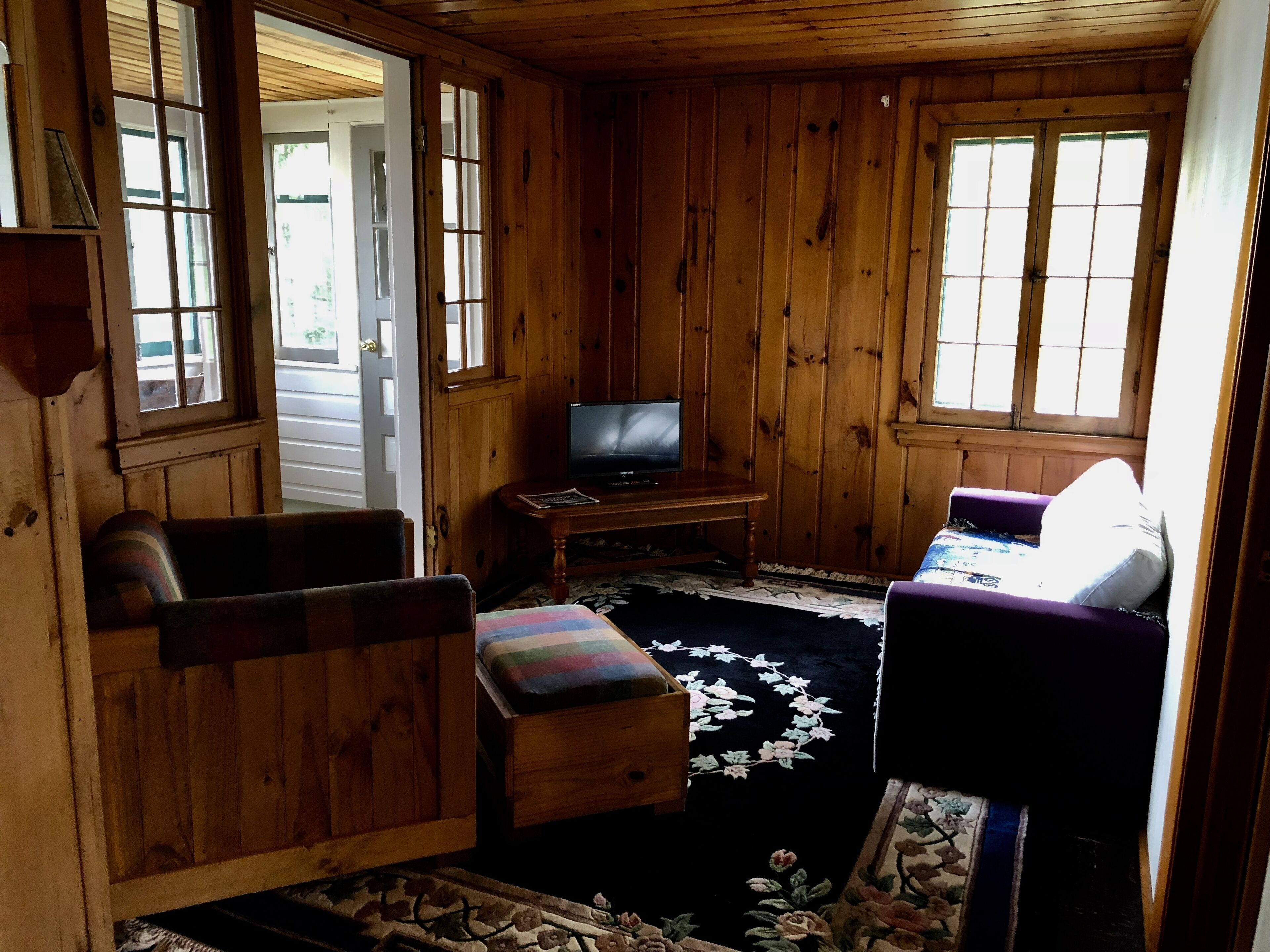 A cozy cabin interior with wooden walls, a television on a wooden table, a cushioned bench, and a patterned rug.