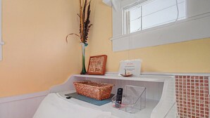 Un baño con lavamanos blanco, una cesta de mimbre, un jarrón con plantas secas, un cuadro enmarco y un letrero que indica acceso a WiFi.