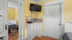 Una habitación con una cama, televisor montado en la pared, una puerta blanca y una pequeña cocina con microondas.