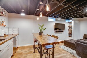A modern kitchen with a wooden table, bar stools, and pendant lights. There is a TV mounted on the wall and a plant on the table.