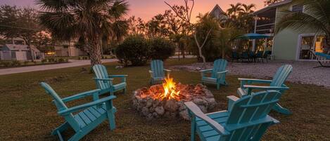 A fire pit with chairs arranged around it, set in a grassy area with palm trees and a house in the background.