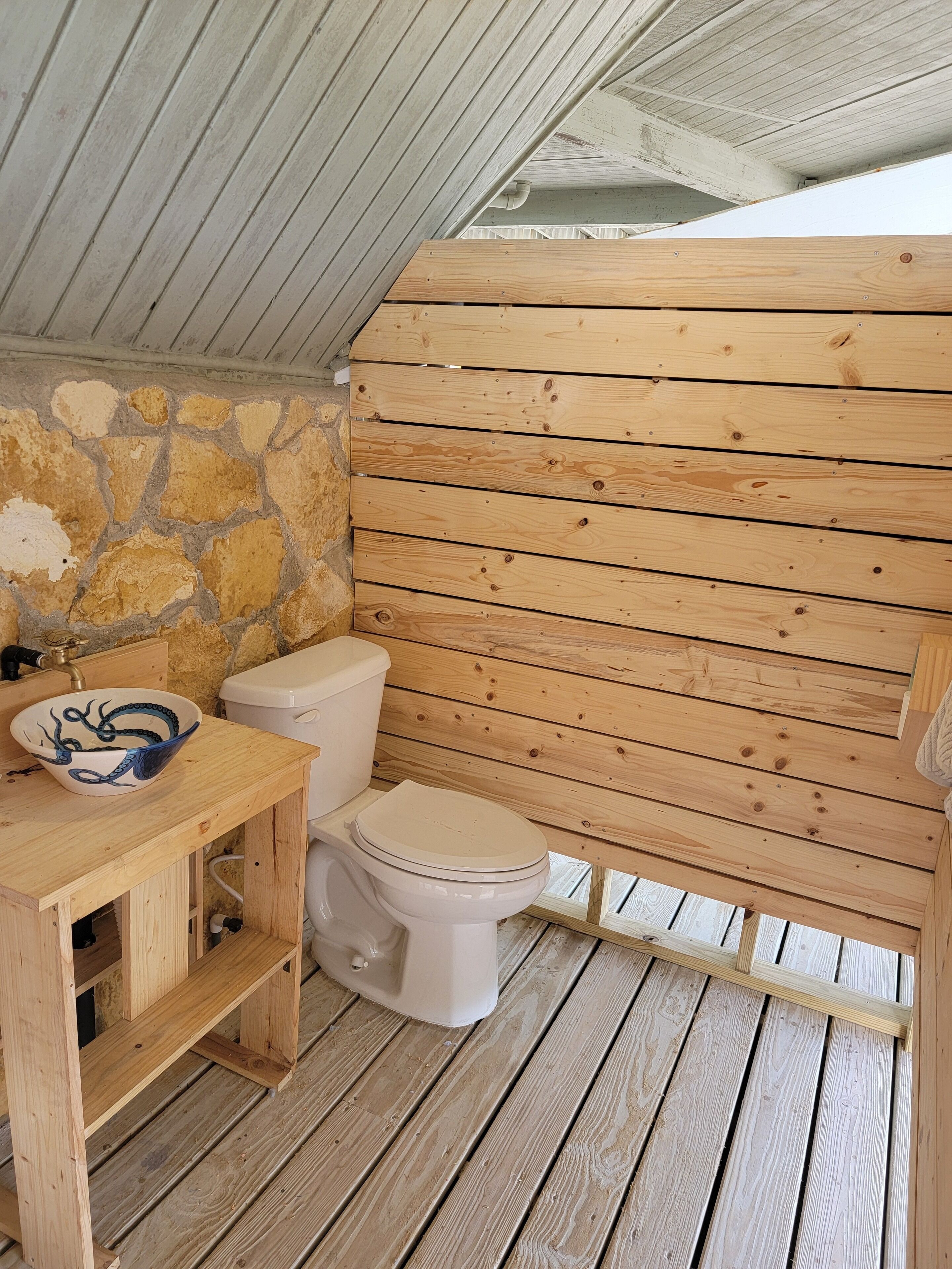 A small, wooden bathroom with a stone wall, a white toilet, and a wooden vanity with a bowl sink.