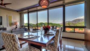 A dining area with a large table set for four, large windows offering a view of a green landscape, and a ceiling fan. - Condo w/ rooftop terrace & ocean views (Playa Hermosa,)