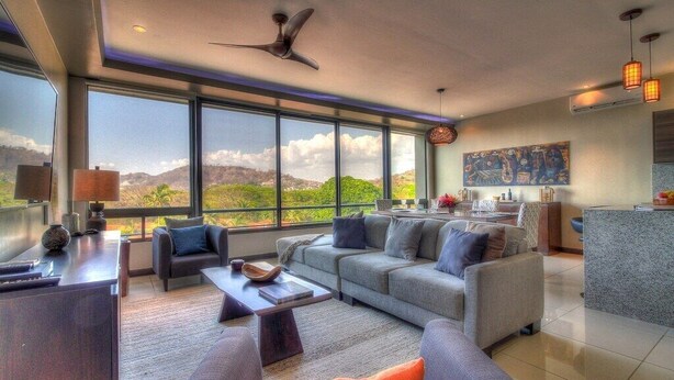 A modern living room with a large window, a ceiling fan, and a kitchen area visible through the window. - Condo w/ rooftop terrace & ocean views (Playa Hermosa,)