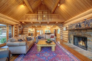 A log cabin interior with a wooden ceiling, a fireplace, a sofa, a coffee table, and a patterned rug.