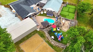 An aerial view of a residential property with a swimming pool, a fenced play area, and a large house with a sloped roof.