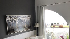 Uma sala de estar com um sofá bege, uma mesa de centro de vidro e um quadro de árvores na parede.