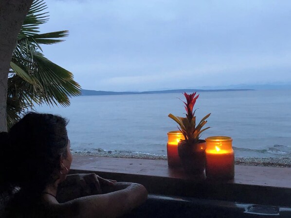 A person seated by a window overlooking a beach with candles on the windowsill. - Oceanside Honeymoon Suite (Qualicum Beach)