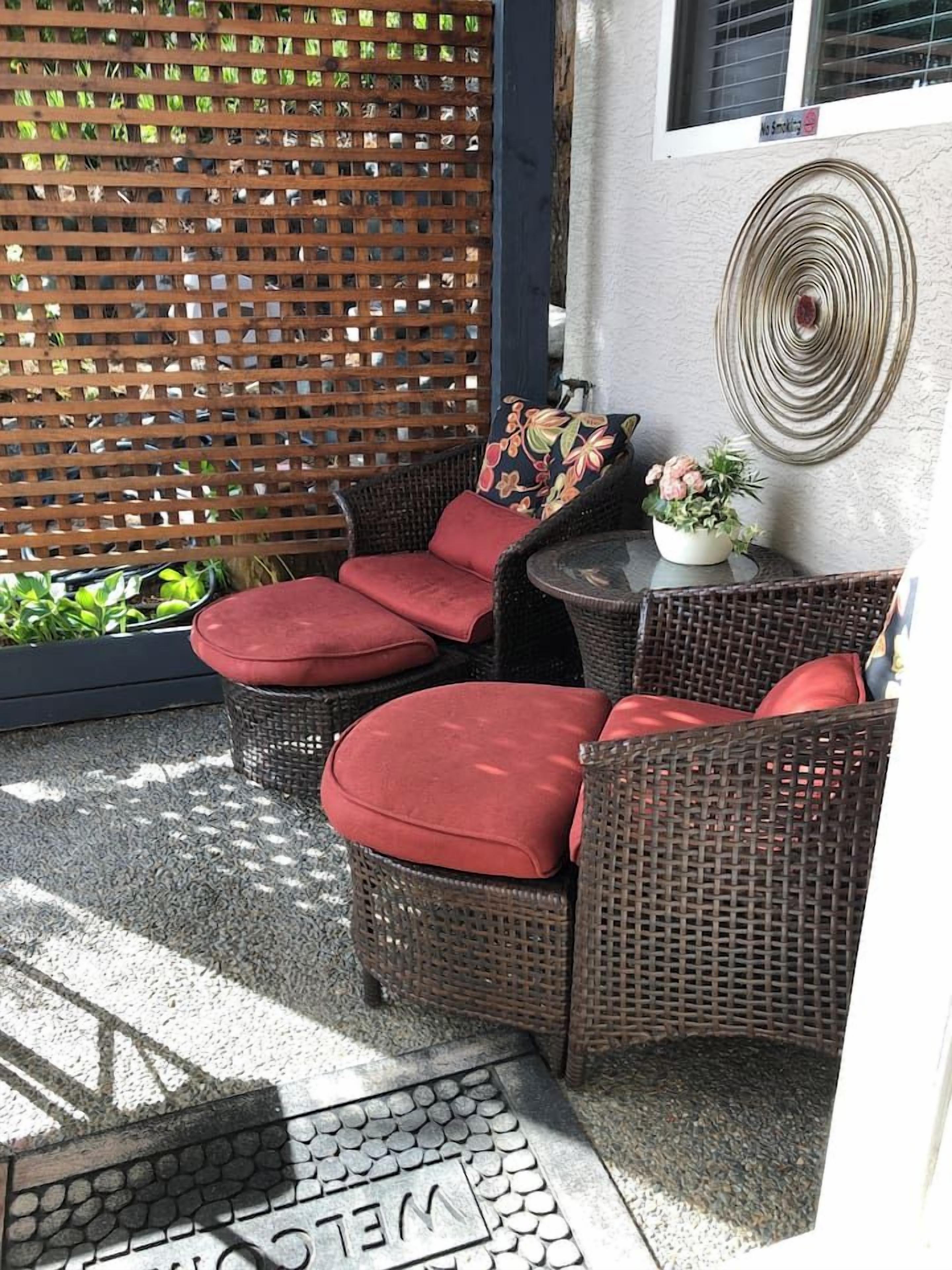 A patio area with wicker furniture and red cushions, a small table, and a decorative wall piece.
