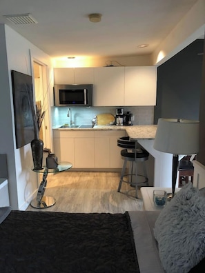 A compact kitchen with a microwave, sink, and bar stools. A flat-screen TV is mounted on the wall, and there is a small seating area with a lamp and a fluffy pillow. - Oceanside Honeymoon Suite (Qualicum Beach)