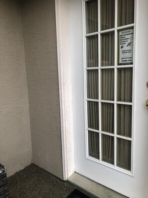 Puerta blanca con un panel de vidrio y un letrero que indica mantener la puerta cerrada y usar zapatos para evitar pulgas de arena y mosquitos.