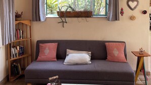A cozy living room with a sofa, a table with decorative items, a bookshelf, and a window with curtains.