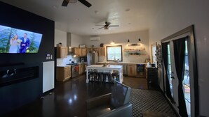 A modern kitchen with wooden cabinets, a dining area with a table and chairs, and a TV displaying a wedding scene. - New Waterfront Flowage to Full Rec Lake Home On UTV Sled Trails Sleeps 10 (Mountain)