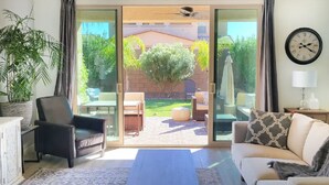 Una sala de estar con sofá, sillón y mesa de centro. Hay un ventanal corredero que da a un patio exterior con césped y sillas.