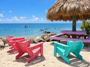 A beach with a thatched-roof structure, a wooden table, and two colorful chairs. - Beautiful Updated Kawama Townhouse.  Marina, boat ramp, lagoon and pools. (Key Largo)