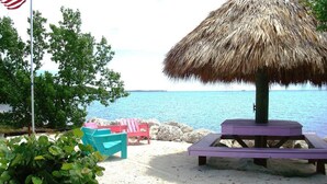A beach scene with a thatched umbrella, a flag, and beach chairs. - Beautiful Updated Kawama Townhouse.  Marina, boat ramp, lagoon and pools. (Key Largo)