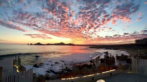 A beachside view with a sunset, a wooden fence, and a fire pit area. - Spectacular View and Luxury Condo - Pet Friendly - Weekly & Monthly discounts  (Cabo San Lucas)