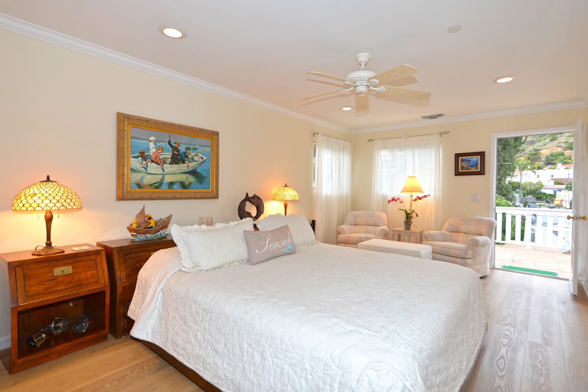 A bedroom with a large bed, two nightstands, a ceiling fan, a painting, and a view of the outdoors.