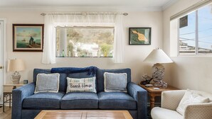 A living room with a blue sofa, a wooden coffee table, and a white armchair. There are two lamps on side tables, a painting on the wall, and a window with a view of buildings. - 220W: Lovely Avalon home + walking distance to the beach, restaurants and more. (Avalon)