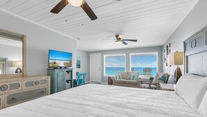 Un espacioso dormitorio con una cama grande, ventilador de techo, televisor y vistas al océano desde las ventanas.
