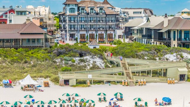 Un hotel frente a la playa con una torre destacada, un paseo marítimo que conduce a la orilla y numerosas sombrillas de playa.