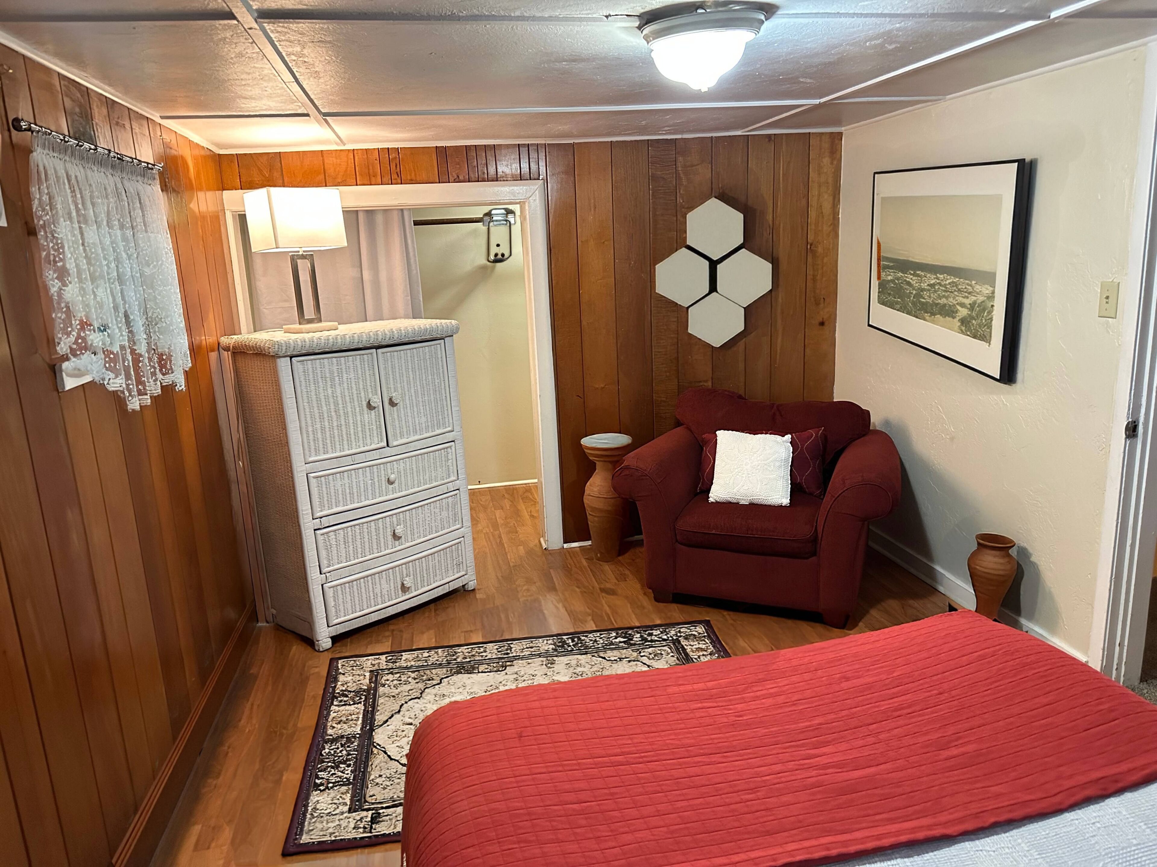 A room with wooden paneling, a white dresser, a red sofa, a bed with a red blanket, a hexagonal wall decoration, and a framed picture on the wall.