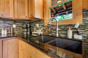 A kitchen with wooden cabinets, a black granite countertop, a stainless steel sink, and a window with a view of greenery. - Arrow Ridge at Hocking Hills (Sugar Grove)
