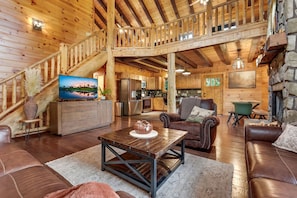 A spacious living area with a wooden staircase, a dining table, leather sofas, and a kitchen in the background. - Arrow Ridge at Hocking Hills (Sugar Grove)