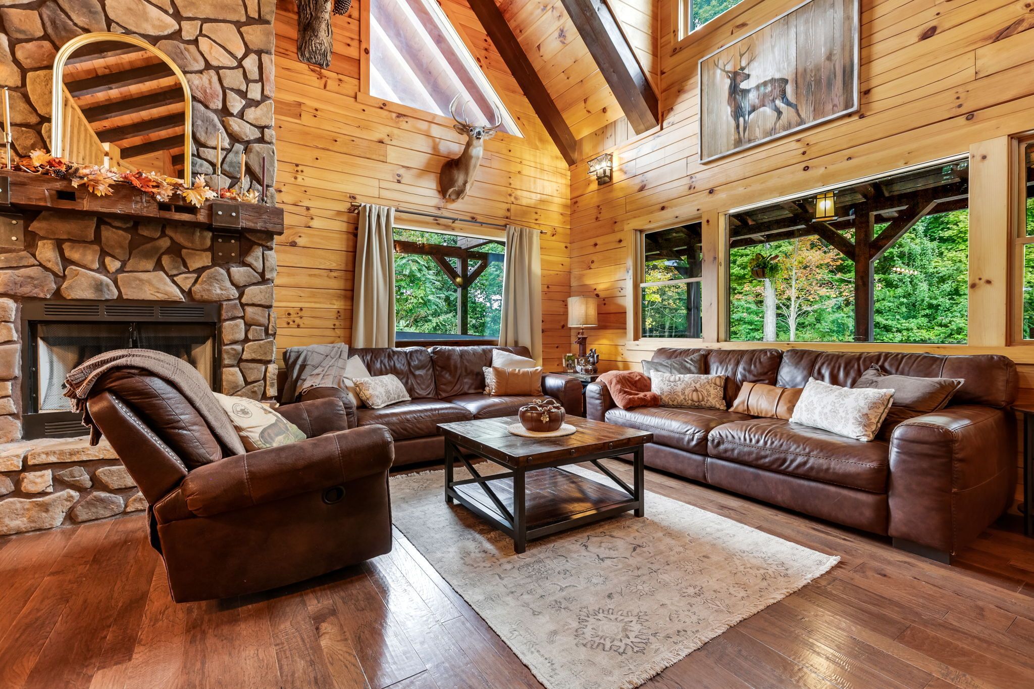 L’intérieur d’une cabane en rondins est agrémenté d’une cheminée en pierre, de canapés en cuir et de grandes fenêtres qui offrent une vue sur la forêt.