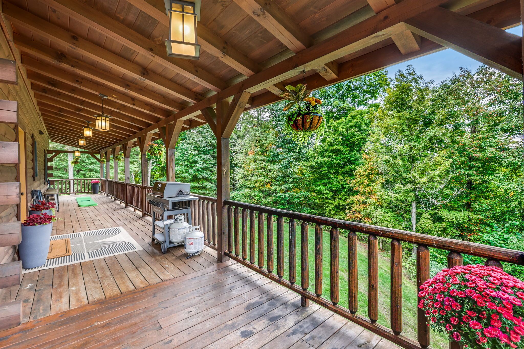 A covered wooden deck with a barbecue grill, outdoor seating, and a view of a forest.