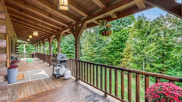 A covered wooden deck with a barbecue grill, outdoor seating, and a view of a forest.